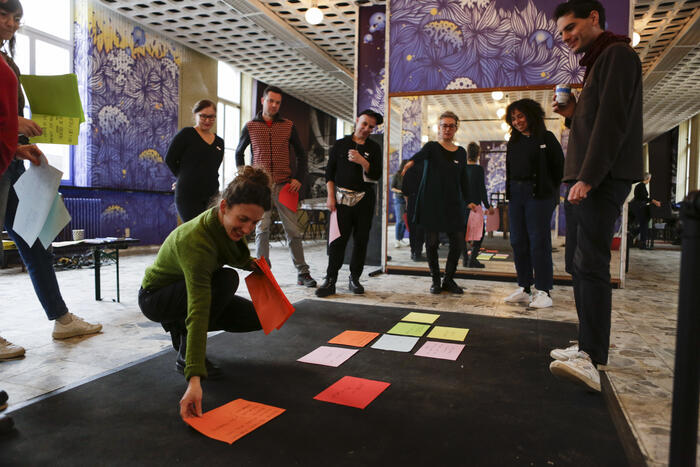 Copy Paste participants circle around a carpet on which are display sheets of colors with handwrittend sentences on them.